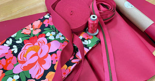A close-up of pink crafting supplies arranged on a wooden surface, including a floral fabric bag, pink ribbon, pink thread on a spool, and a roll of pink material.